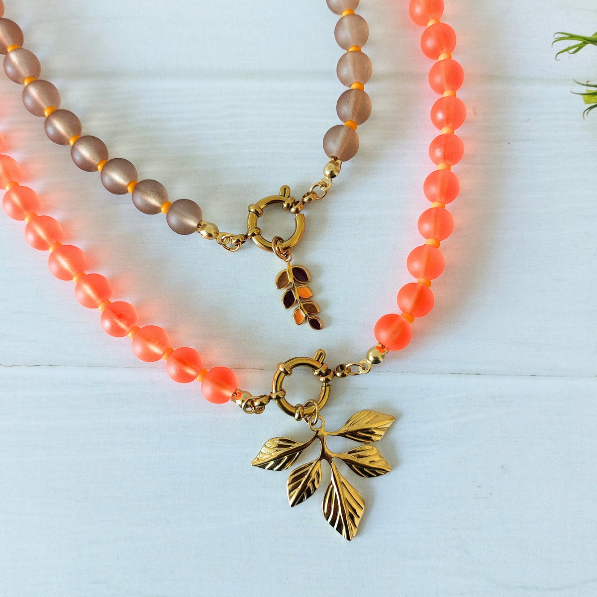 Two beaded necklaces displayed on a  wooden surface. One necklace features translucent peach-orange beads with a gold leaf pendant, and the other features smoky brown translucent beads with a gold clasp and charm.