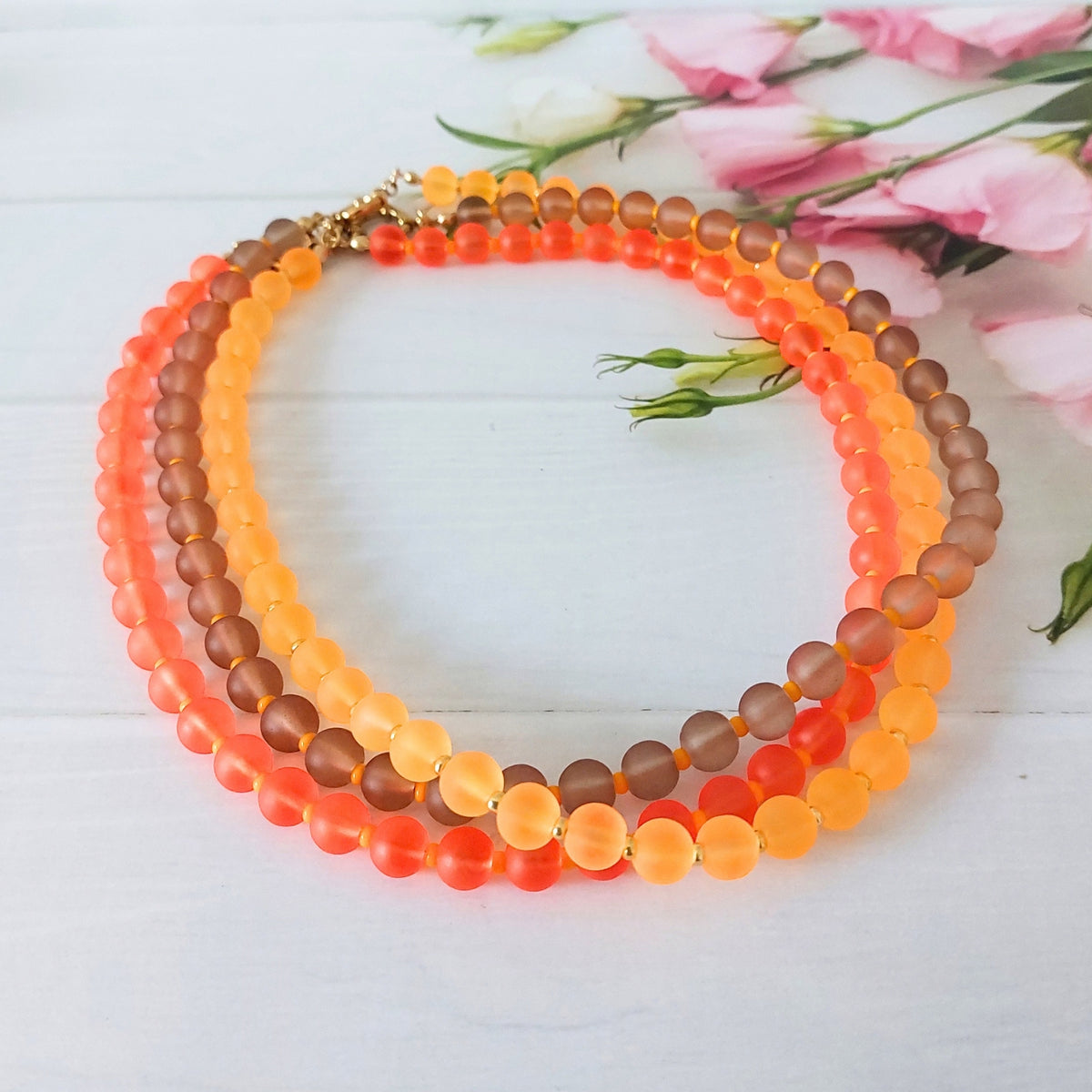 A collection of 3 beaded necklaces with frosted round beads in orange, coral, and brown tones, arranged in a circle on a white wooden surface with pink flowers in the background.