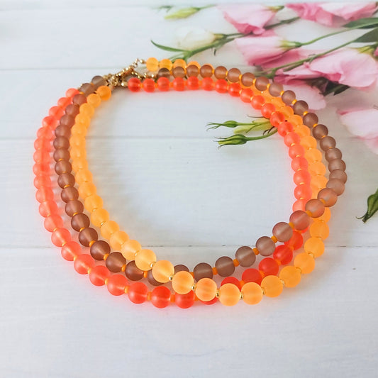 A collection of 3 beaded necklaces with frosted round beads in orange, coral, and brown tones, arranged in a circle on a white wooden surface with pink flowers in the background.