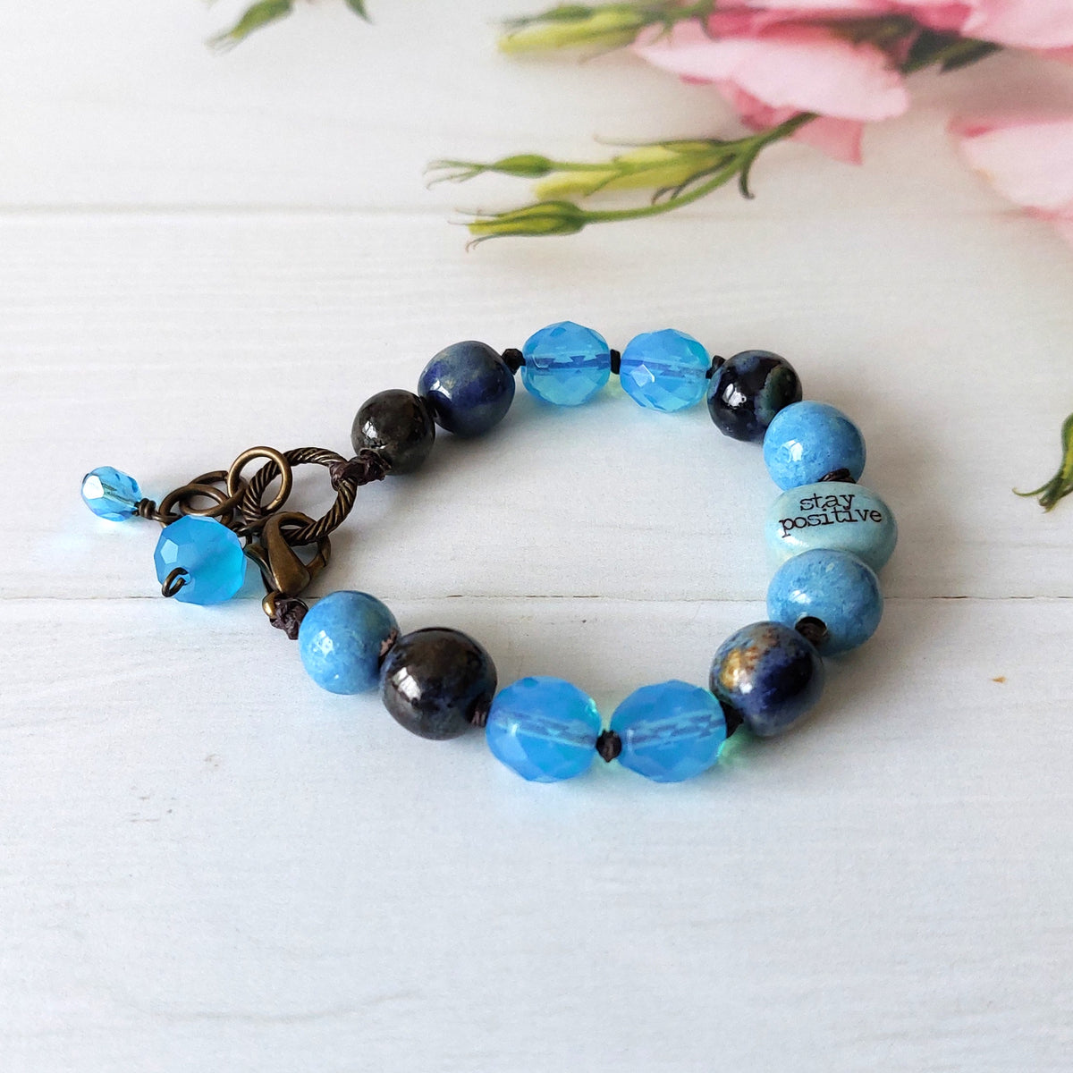 A beaded bracelet with light blue, dark blue, and dark greybeads, featuring a bronze-toned clasp and a pale blue bead with the words 'stay positive' printed on it. The bracelet is displayed on a white wooden surface with soft pink flowers in the background.