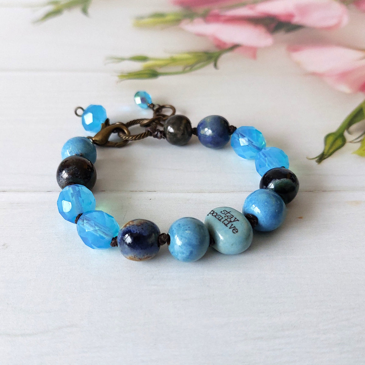 A beaded bracelet with blue, navy, and grey beads, featuring a rustic bronze clasp and a light blue bead printed with 'stay positive'. The bracelet is displayed on a white wooden surface with soft pink flowers in the background.