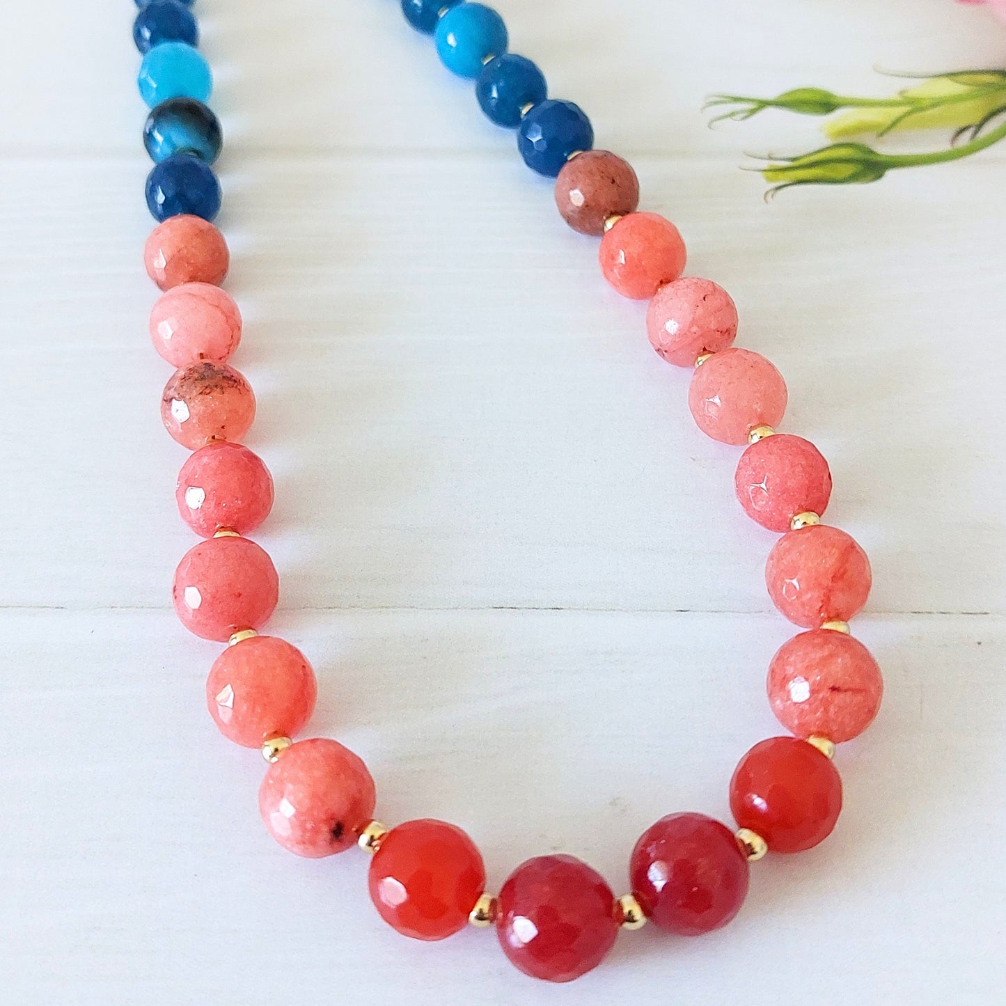 A jade beaded necklace with an ombre style of translucent coral-pink beads into deep red beads into blue/turquoise beads, separated by small gold-plated stainless steel spacer beads, displayed on a white wooden surface.