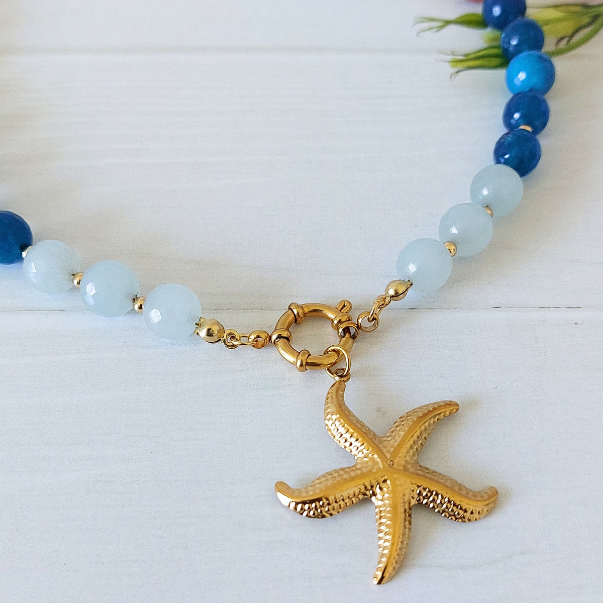 A jade beaded necklace with light blue and dark blue round beads, gold-plated stainless steel links, and a textured gold-plated stainless steel starfish pendant, displayed on a white wooden surface.