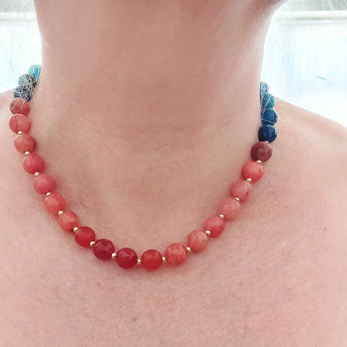 A jade beaded necklace worn on a person's neck, featuring round coral-pink and red beads separated by small gold-plated stainless steel spacers, with blue beads at the back, shown against skin.