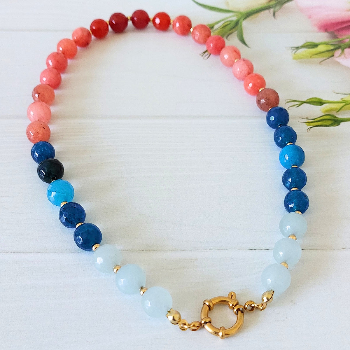 A beaded necklace with alternating sections of coral, red, dark blue, light blue, and pale aqua beads, finished with a gold-plated stainless steel statement spring clasp, displayed on a white wooden surface with pink flowers in the background.