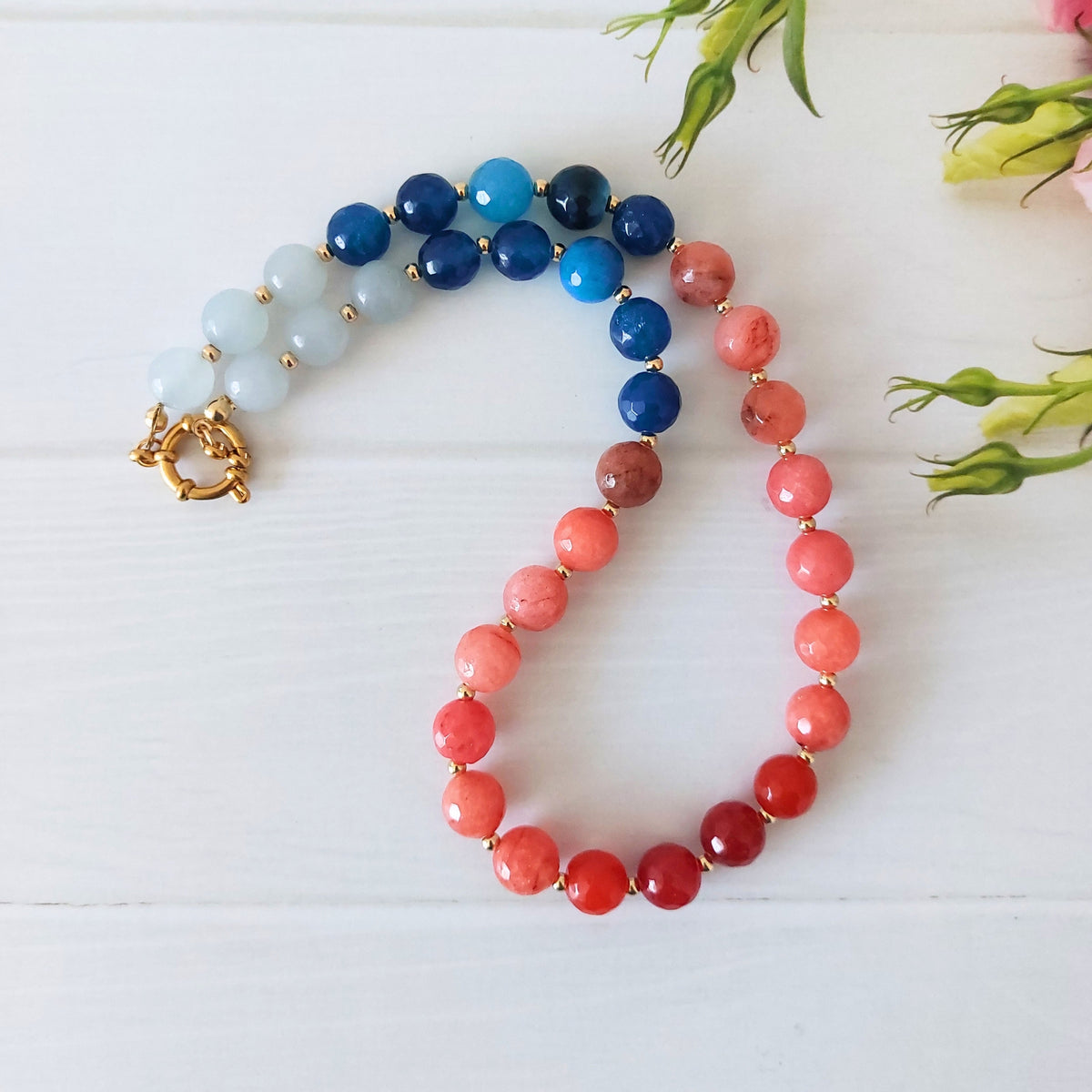 A beaded necklace arranged in a loop on a white wood surface, featuring pale icy blue beads, deep blue beads, coral-pink beads, and red beads, separated by small gold spacer beads and finished with a gold-plated stainless steel spring clasp.