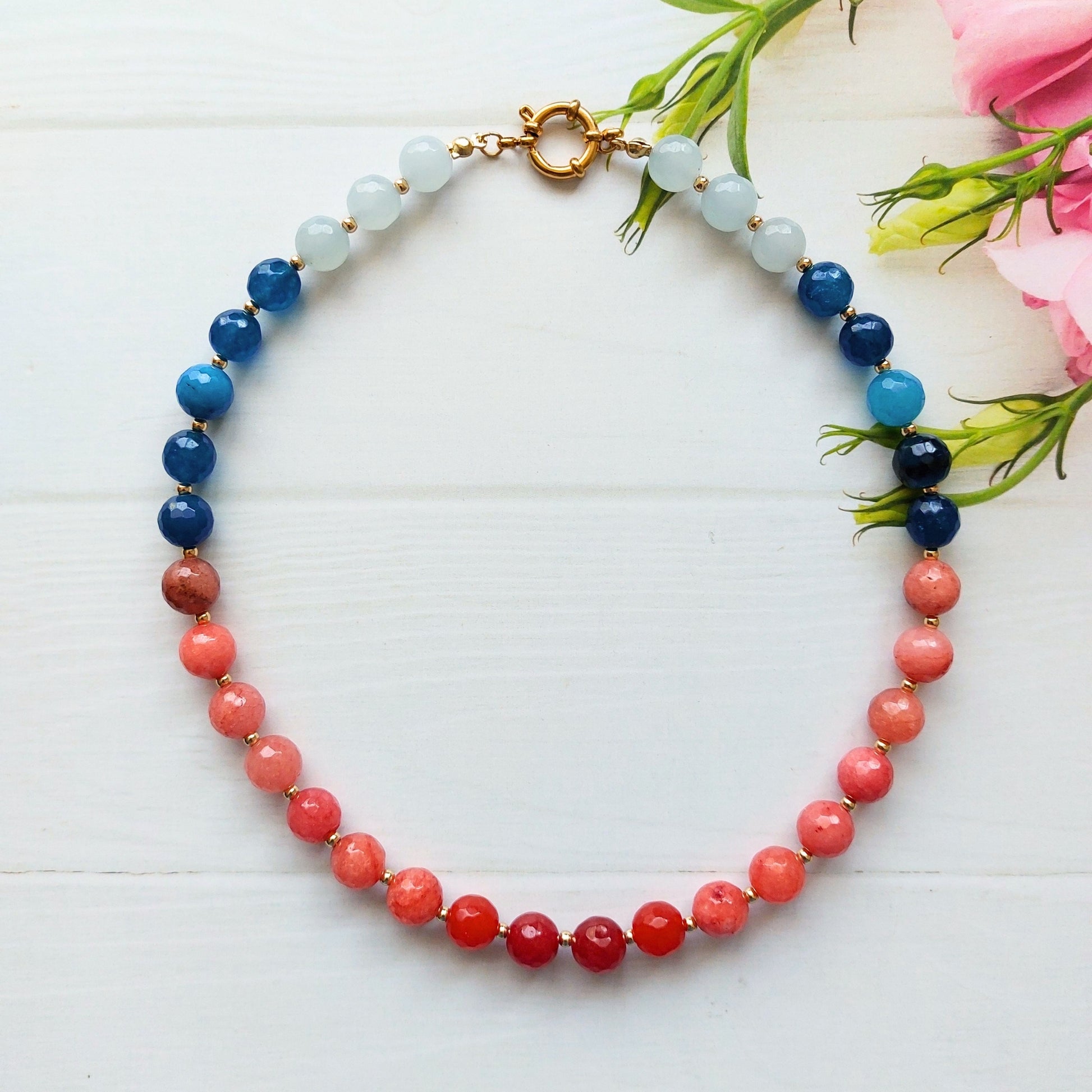 A beaded necklace arranged in a circle on a white wooden surface, with pink flowers placed in the top-right corner. The necklace features a gradient of round beads transitioning from pale blue at the top, through blue, to coral pink and deep red at the bottom, finished with a gold-plated stainless steel clasp.