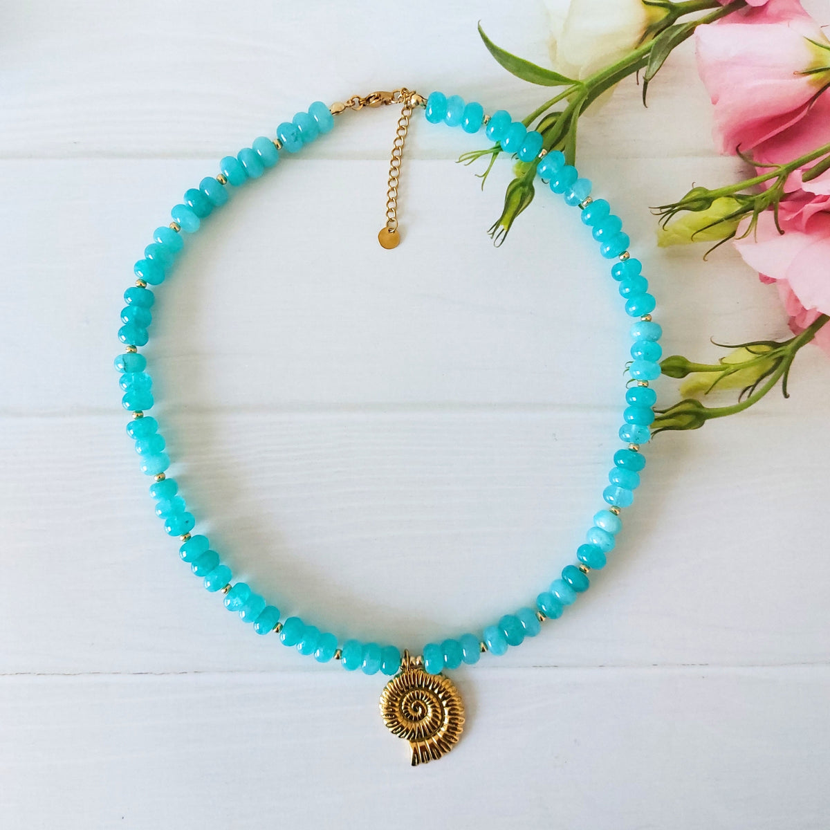 A turquoise beaded jade gemstone necklace with a gold-plated stainless steel spiral shell pendant, displayed on a white wooden background next to pink flowers.