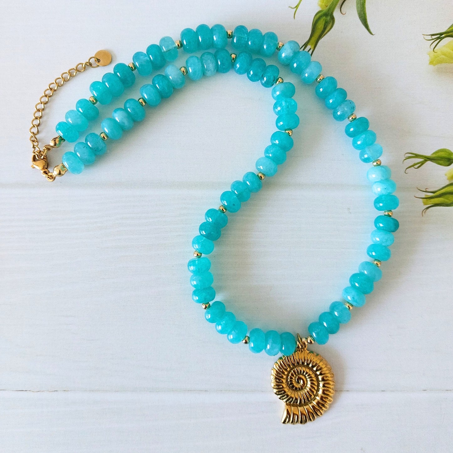 A turquoise jade beaded necklace with a gold-plated stainless steel spiral shell pendant, displayed on a white wooden surface with small green leaves in the background.