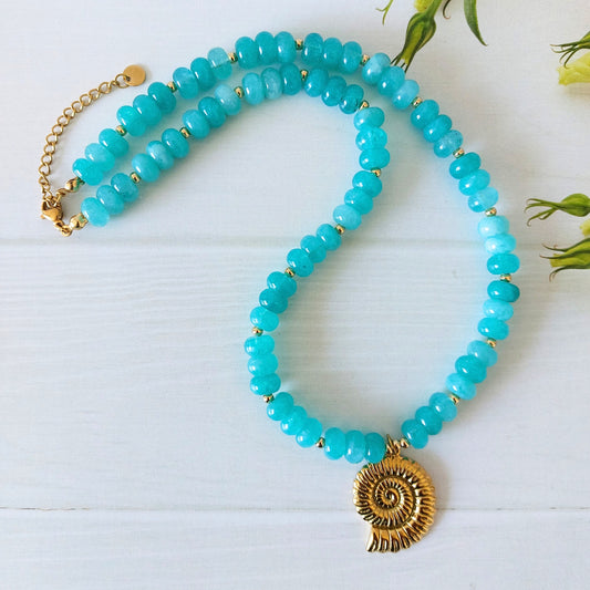 A turquoise jade beaded necklace with a gold-plated stainless steel spiral shell pendant, displayed on a white wooden surface with small green leaves in the background.