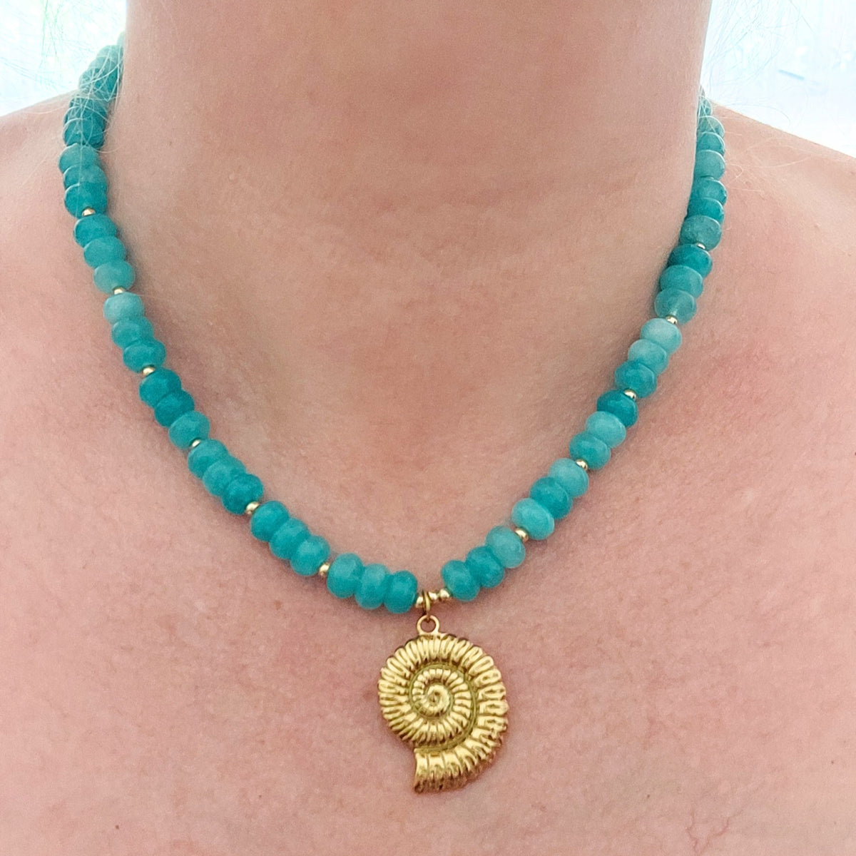 A close-up of a person's neck and upper chest wearing a turquoise jade beaded necklace with a gold-plated stainless steel spiral shell pendant.