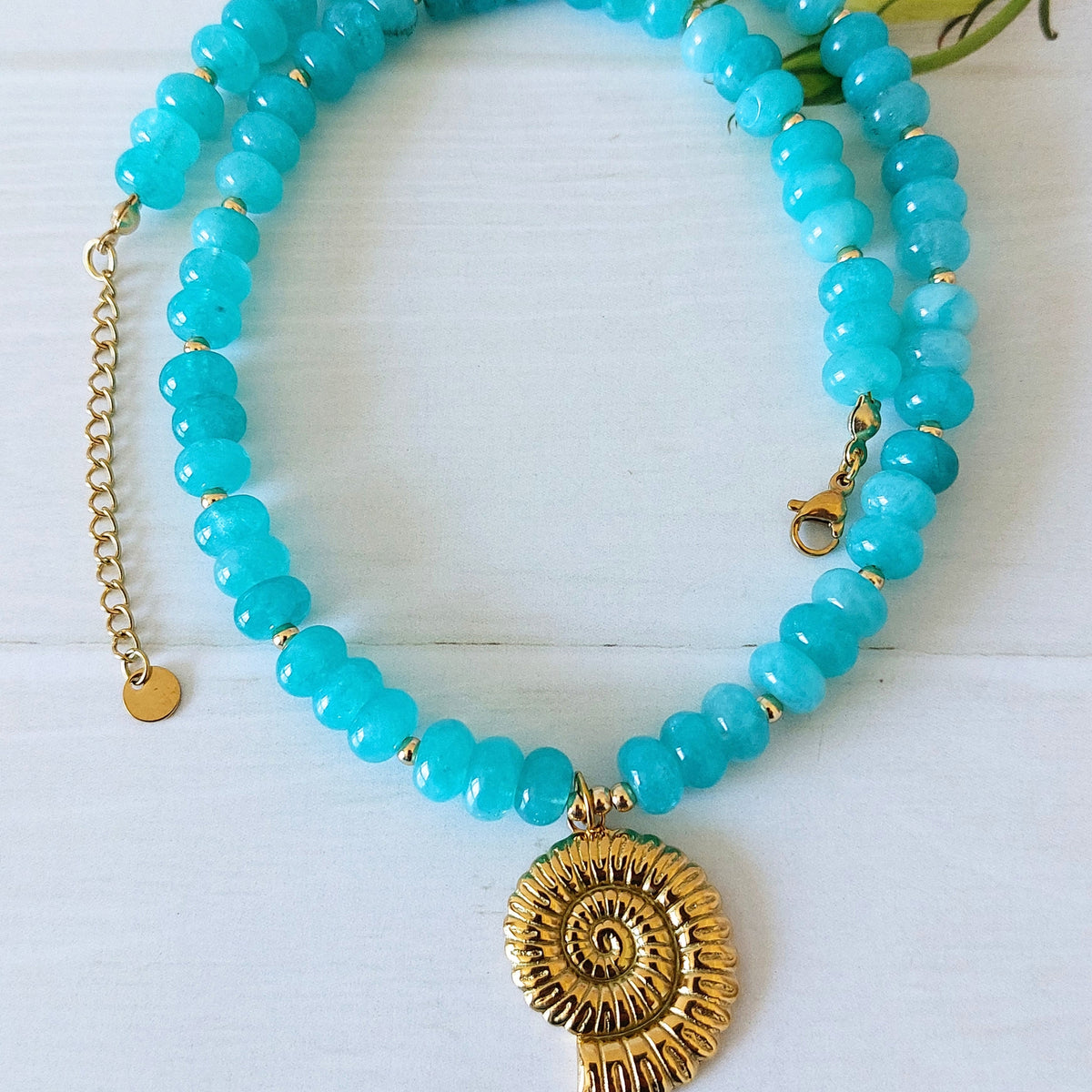 A turquoise jade beaded necklace with a gold-plated stainless steel spiral shell pendant, displayed on a white wooden background.