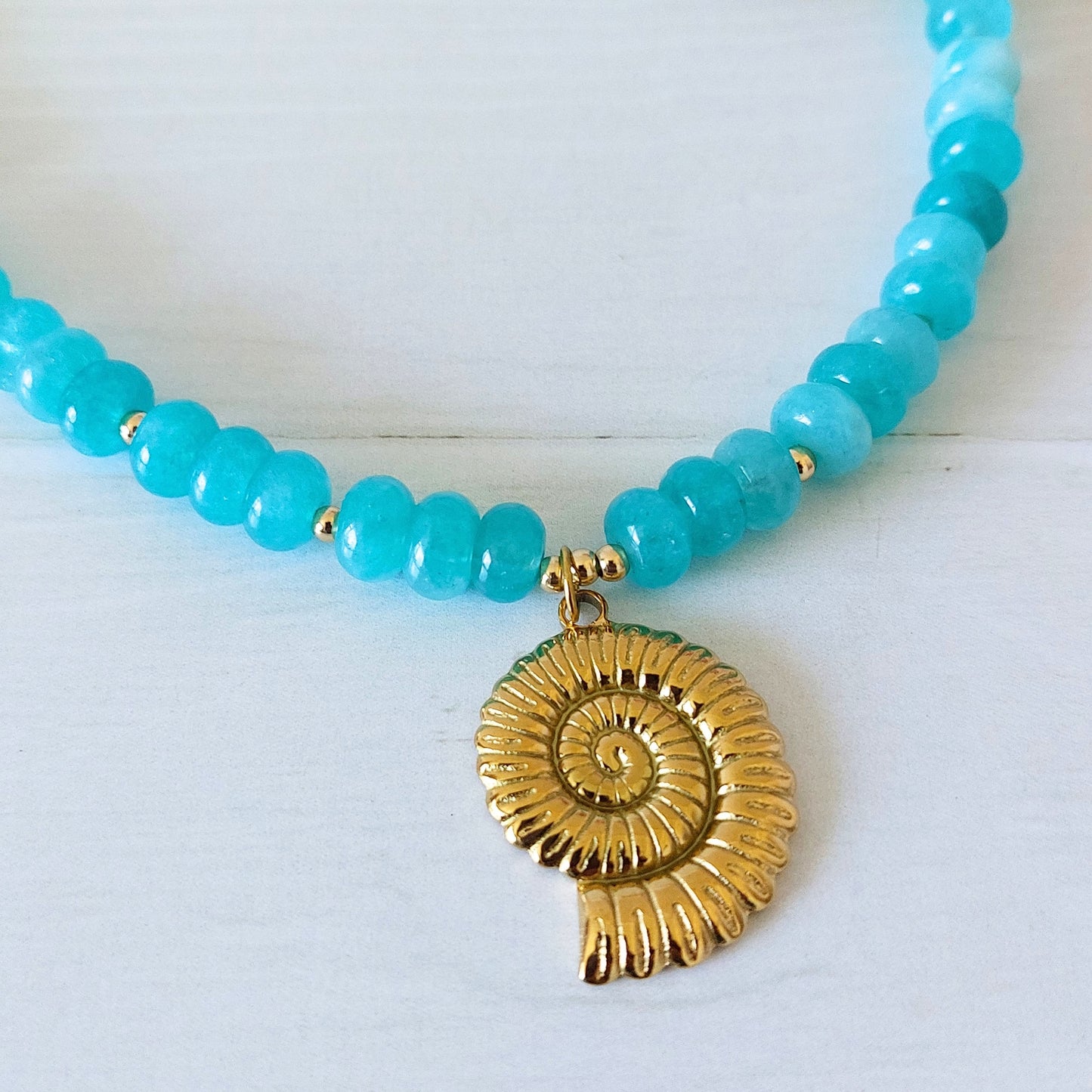 A beaded necklace with turquoise jade rondelle beads and a textured gold-plated stainless steel spiral shell pendant, displayed on a white wooden surface.