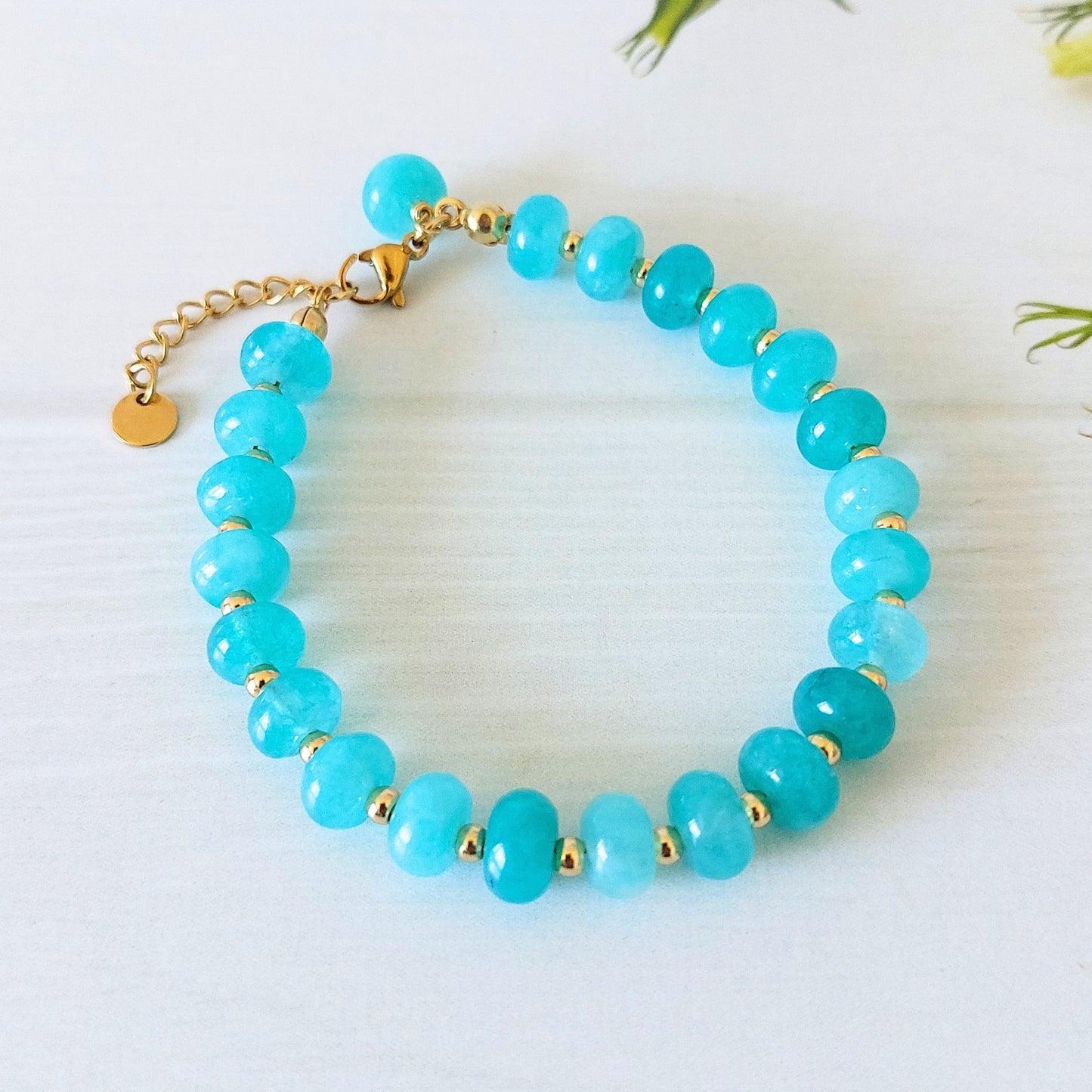 A beaded bracelet made from turquoise-blue jade beads separated by small gold spacer beads, with a gold-plated stainless steel clasp and extender chain on a white surface.