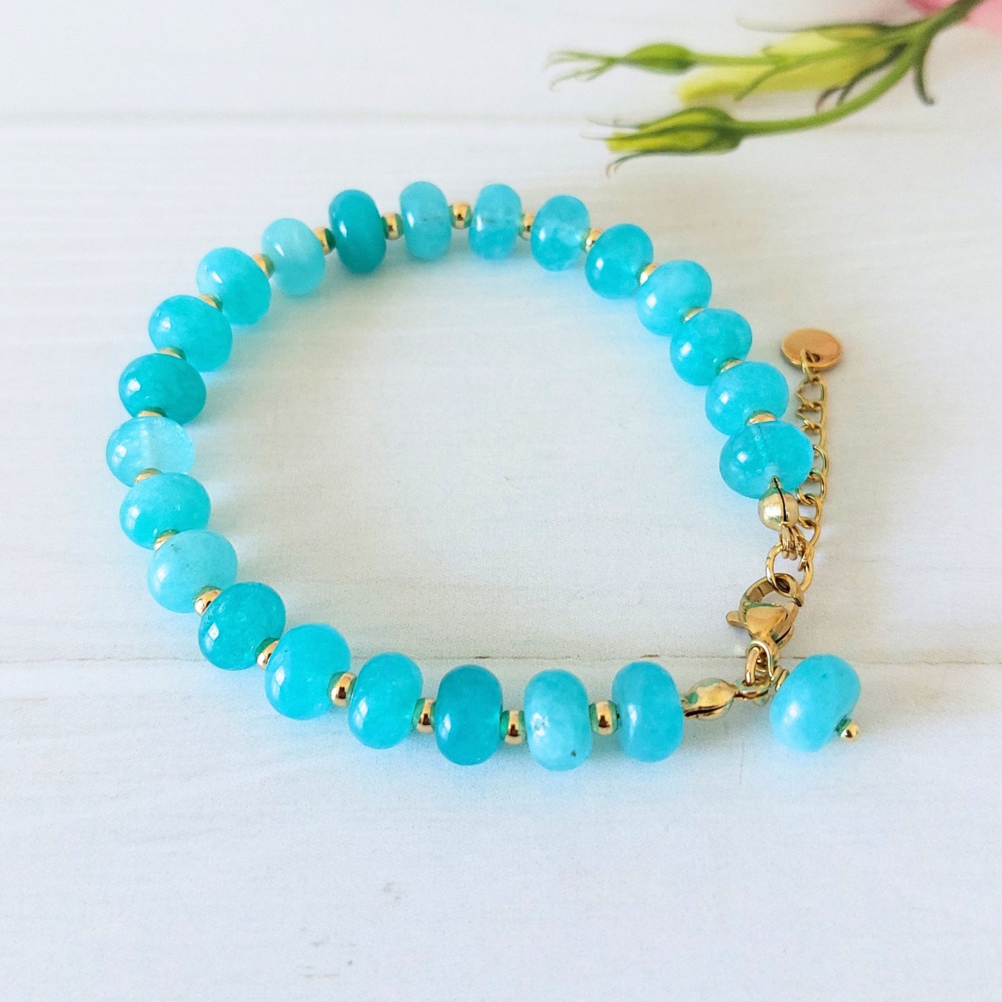 A beaded bracelet made of turquoise-blue rondelle beads separated by small gold spacer beads, with a gold-plated stainless steel lobster clasp and extender chain, photographed on a white wooden surface.