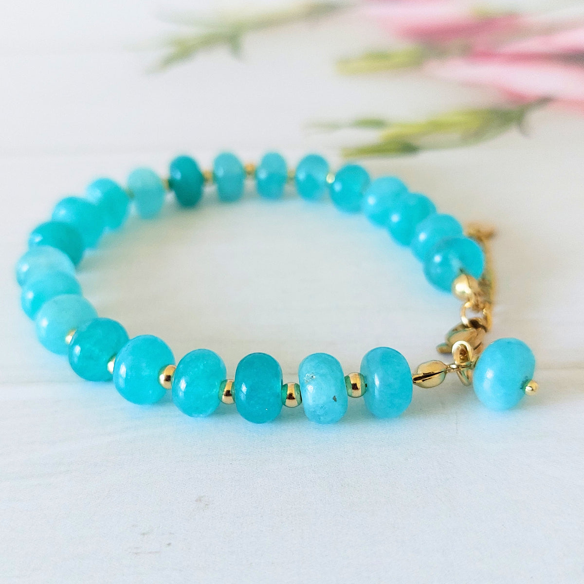 A beaded bracelet made of turquoise-blue gemstone beads separated by small gold spacer beads, with a gold-plated stainless steel clasp and a dangling charm, photographed on a light surface with soft blurred flowers in the background.