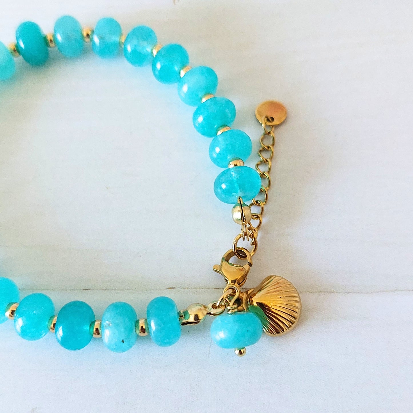 A turquoise beaded bracelet with gold-plated stainless steel spacer beads, a gold lobster clasp, and a gold shell charm, displayed on a white wooden background.