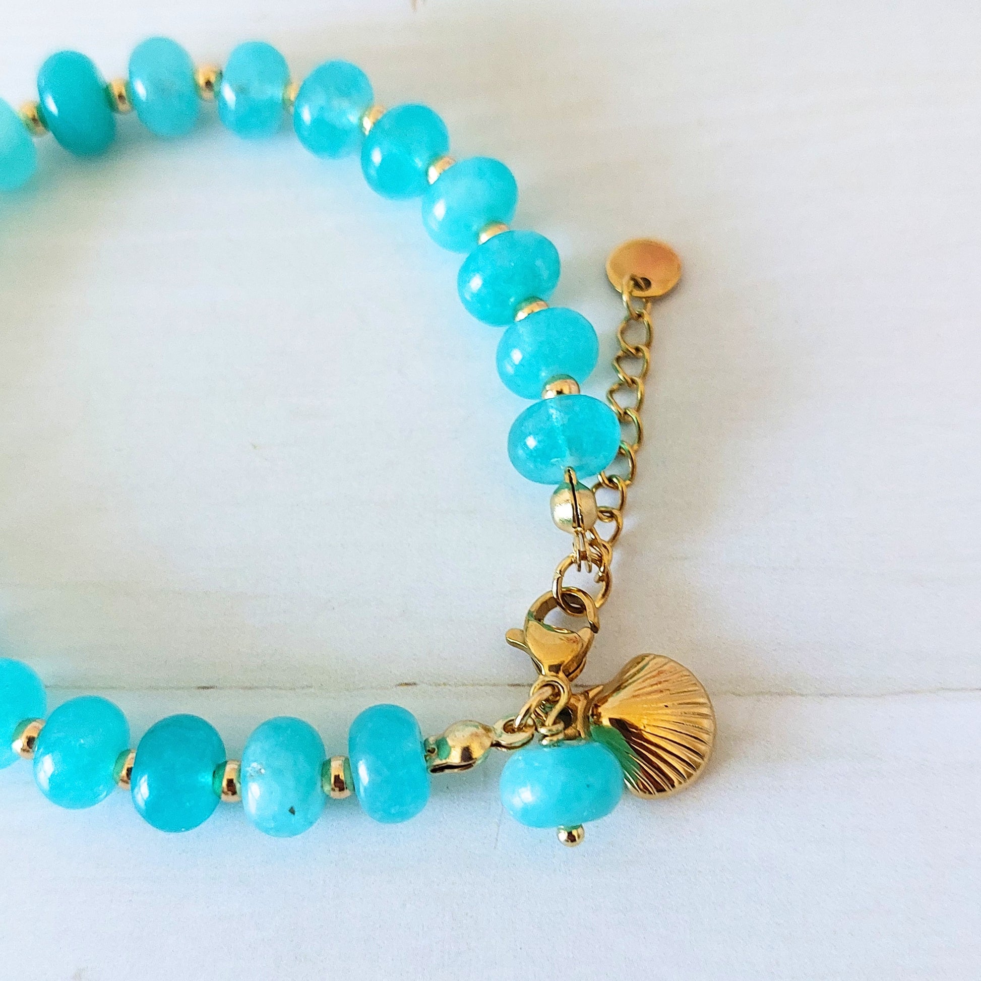 A turquoise beaded bracelet with gold-plated stainless steel spacer beads, a gold lobster clasp, and a gold shell charm, displayed on a white wooden background.