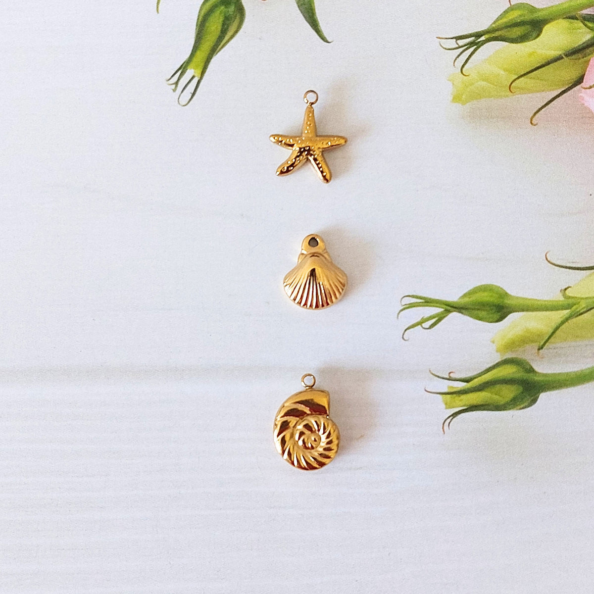 Three small gold-toned jewelry charms shaped like a starfish, a scallop shell, and a spiral shell, arranged vertically on a white background with green flower buds as decorative props.