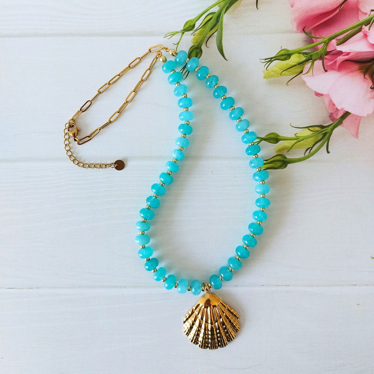 A turquoise jade bead necklace with a gold-plated stainless steel shell pendant, displayed on a white wooden surface next to pink flowers.