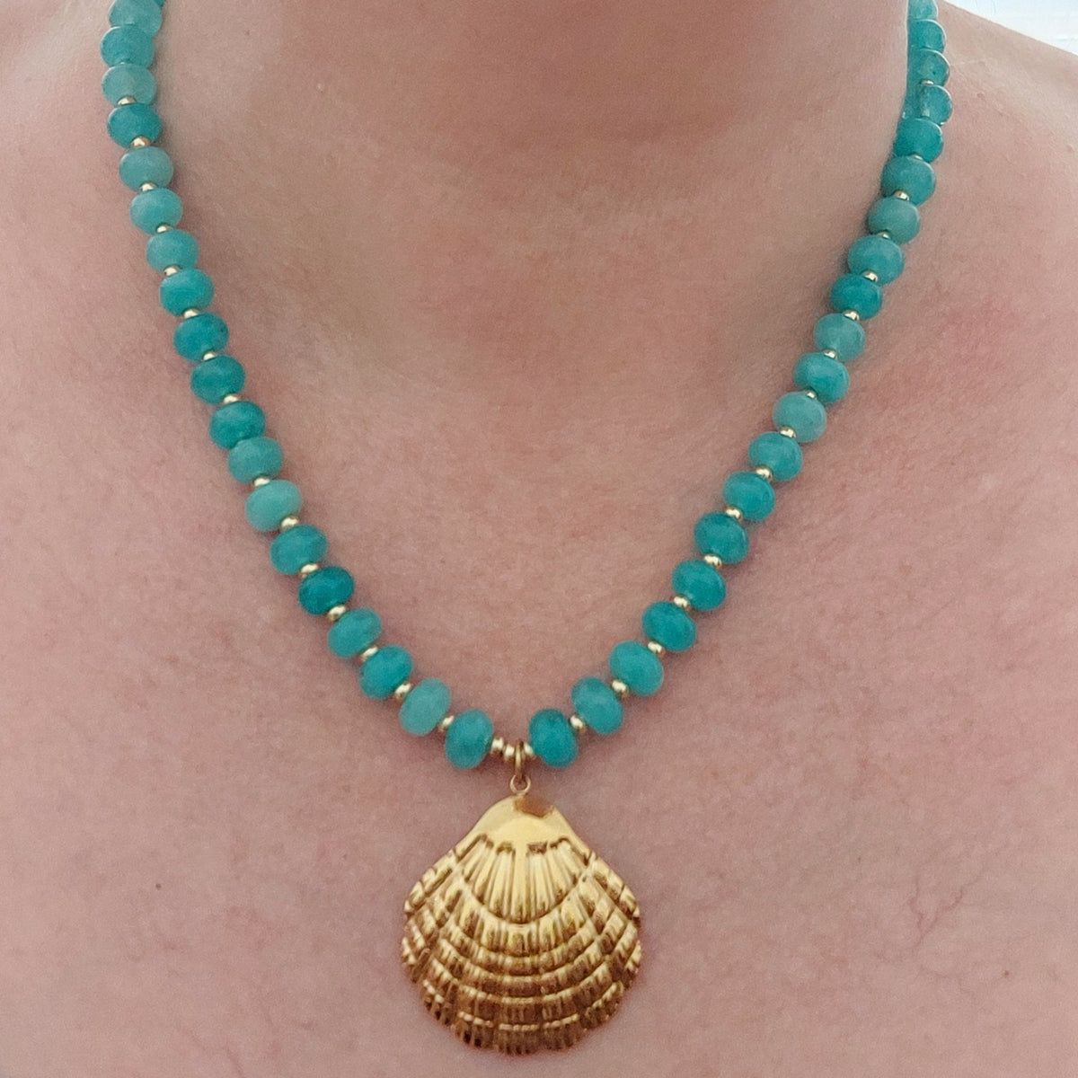 A turquoise jade gemstone necklace with a gold-plated stainless steel shell pendant, shown worn on a person's neck.