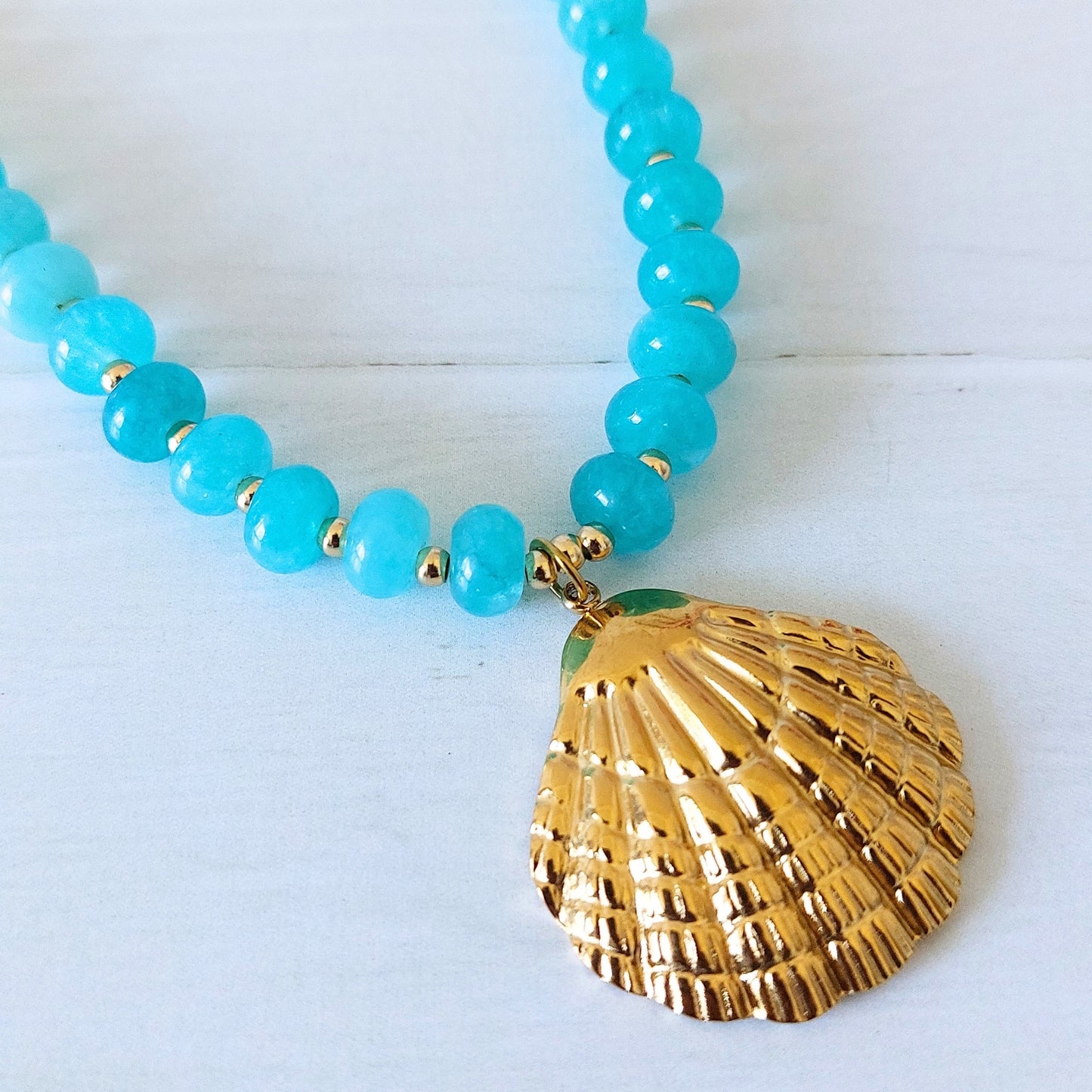 A beaded necklace with bright turquoise-blue jade rondelle beads separated by small gold-plated stainless steel spacer beads, featuring a large textured gold shell pendant, displayed on a light wooden surface.