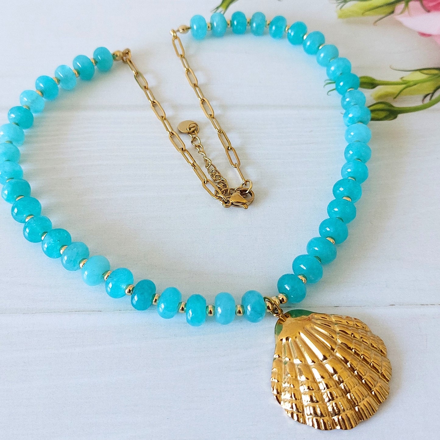A turquoise jade bead necklace with a gold-plated stainless steel shell pendant with a gold-plated paperclip chain finish, displayed on a white wooden surface and small pink flower buds in the corner.