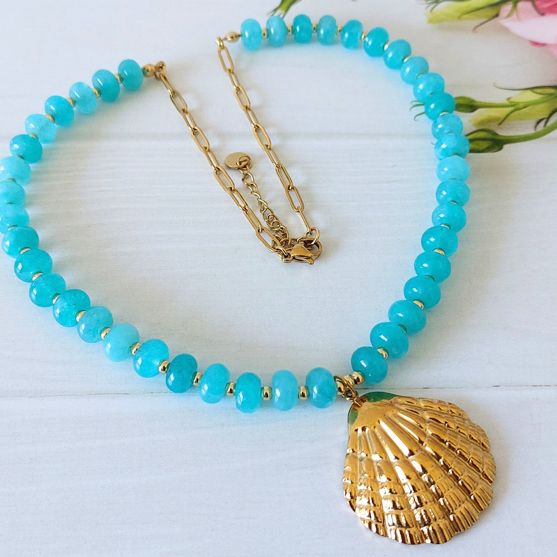 A turquoise jade bead necklace with a gold-plated stainless steel shell pendant with a gold-plated paperclip chain finish, displayed on a white wooden surface and small pink flower buds in the corner.