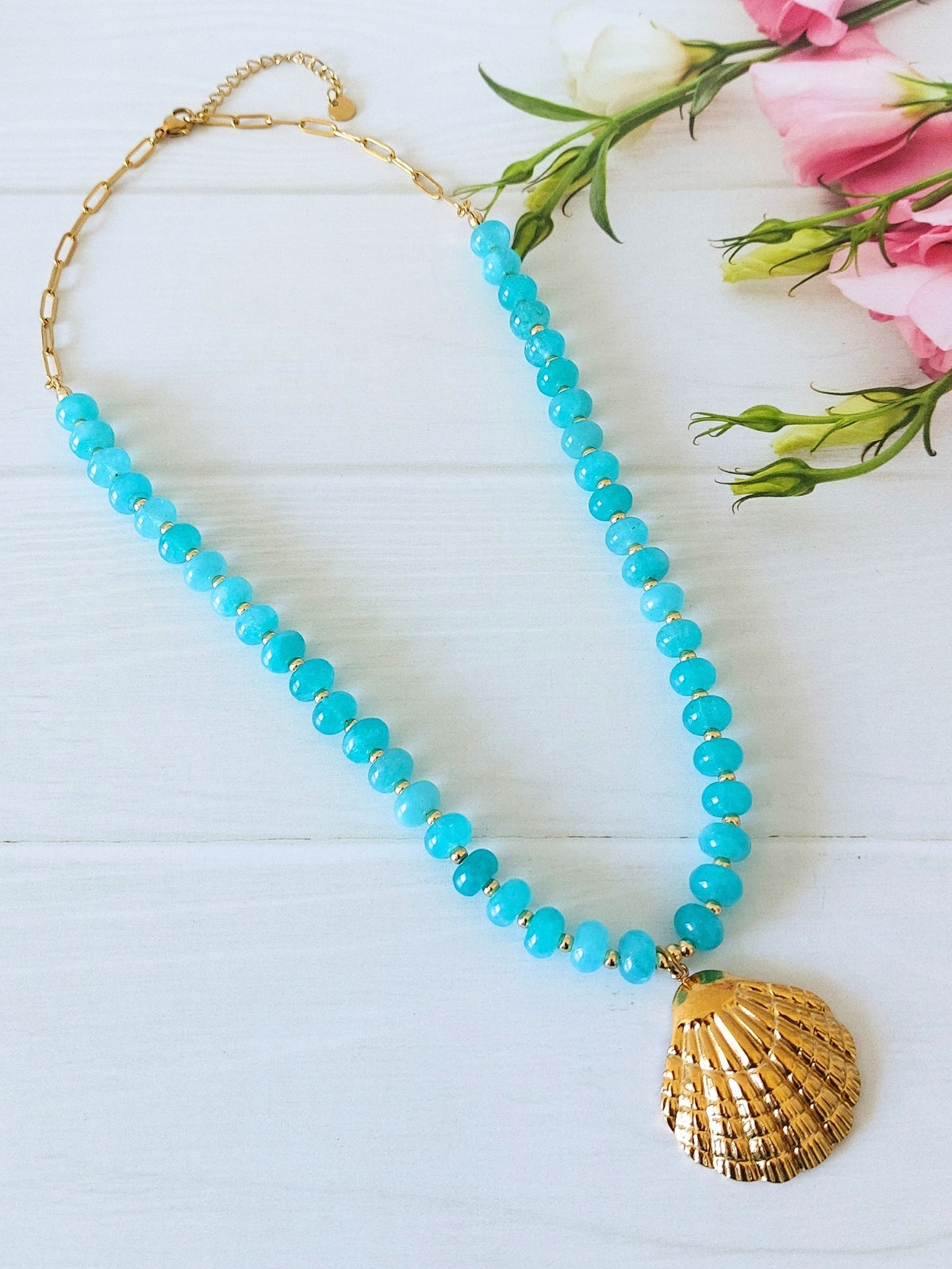 A gold-plated stainless steel paperclip chain and jade bead necklace in turquoise blue beads with a gold-plated seashell pendant, displayed on a white wooden surface next to pink flowers.