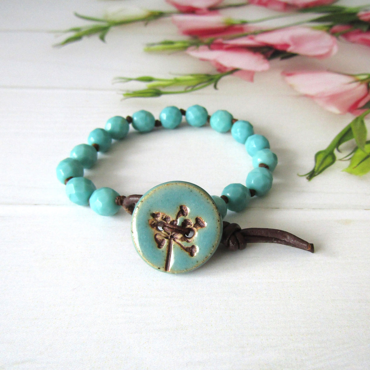 A turquoise beaded bracelet with a round turquoise button featuring an imprint of a seed-head, tied with dark brown cord, displayed on a white wooden surface with pink flowers in the background.