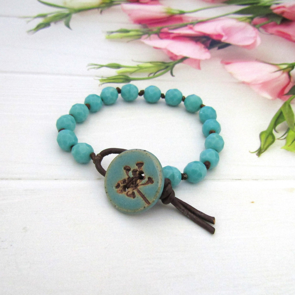 A turquoise bead bracelet with a large round turquoise button featuring a brown seed-head design, knotted with dark brown cord, displayed on a white wooden surface with pink flowers in the background.