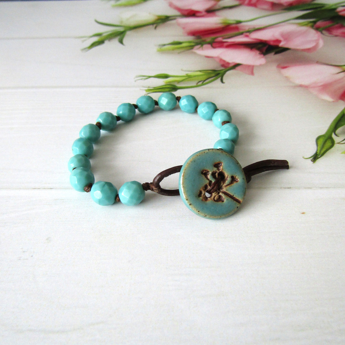 A turquoise beaded bracelet with a large round turquoise button closure featuring a seed-head design, resting on a white wooden surface with pink flowers in the background.