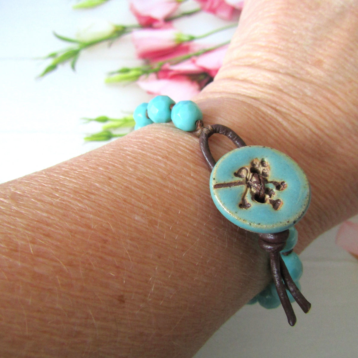 A close-up of a person's wrist wearing a turquoise and dark brown beaded bracelet with a large round turquoise button featuring a seed-head design.