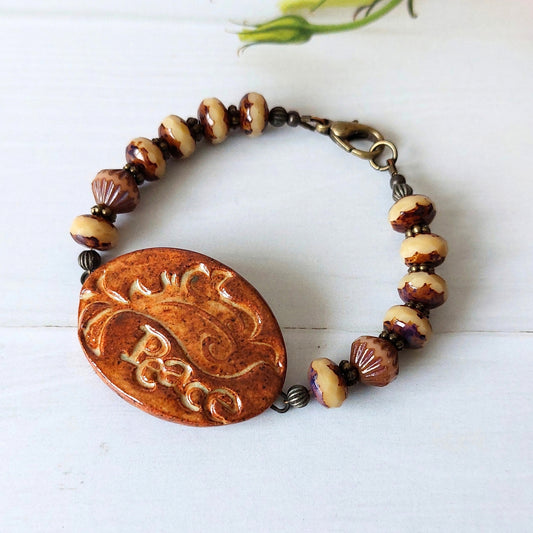 A handmade beaded bracelet with a central ceramic connector with a bird design and the word 'Peace' and brown & cream patterned beads, displayed on a white wooden surface.