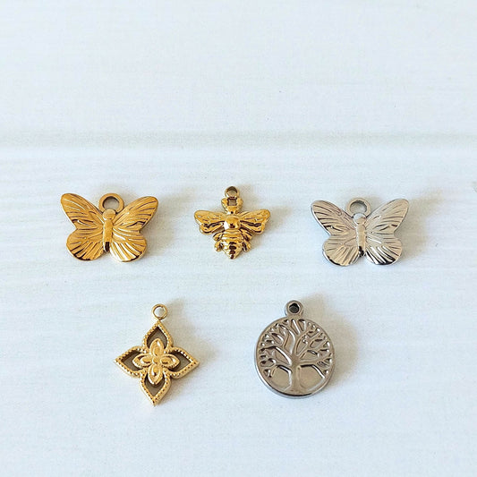 Five charms on a wooden background including a gold-tone and silver-tone butterfly, a gold-tone bee, a gold-tone flower motif and a silver-tone tree of life charm.