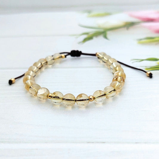 A beaded bracelet featuring soft yellow faceted round Citrine beads with gold metallic accents and a brown adjustable cord, displayed on a white wooden surface with soft pink flowers in the background.