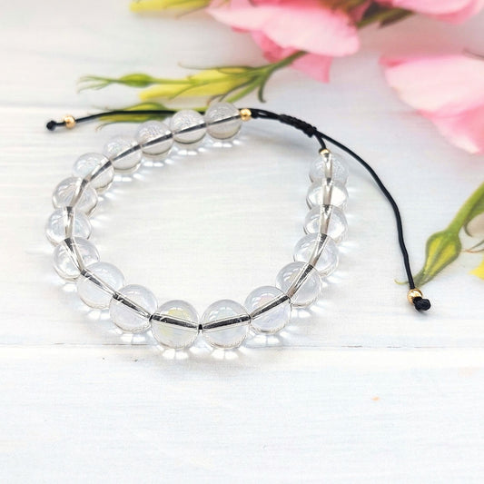 Clear beaded bracelet with black cord,  accented with small gold spacers, arranged on a white wooden surface with pink flowers in the background.