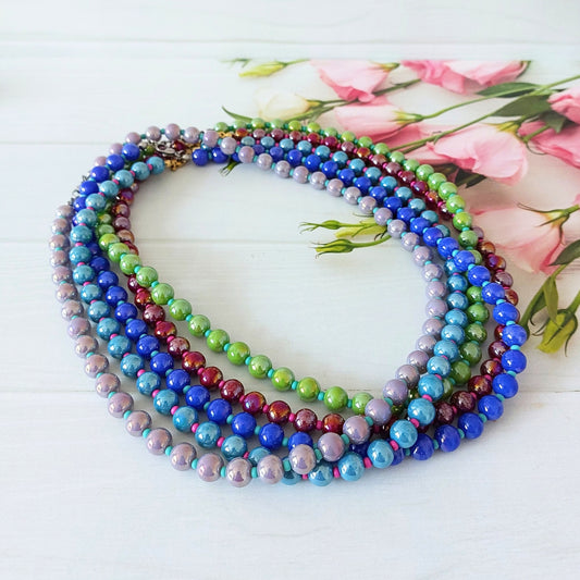 A stack of colourful beaded necklaces with iridescent round beads in blue, purple, green, and red, arranged on a white wooden surface with pink roses in the background.