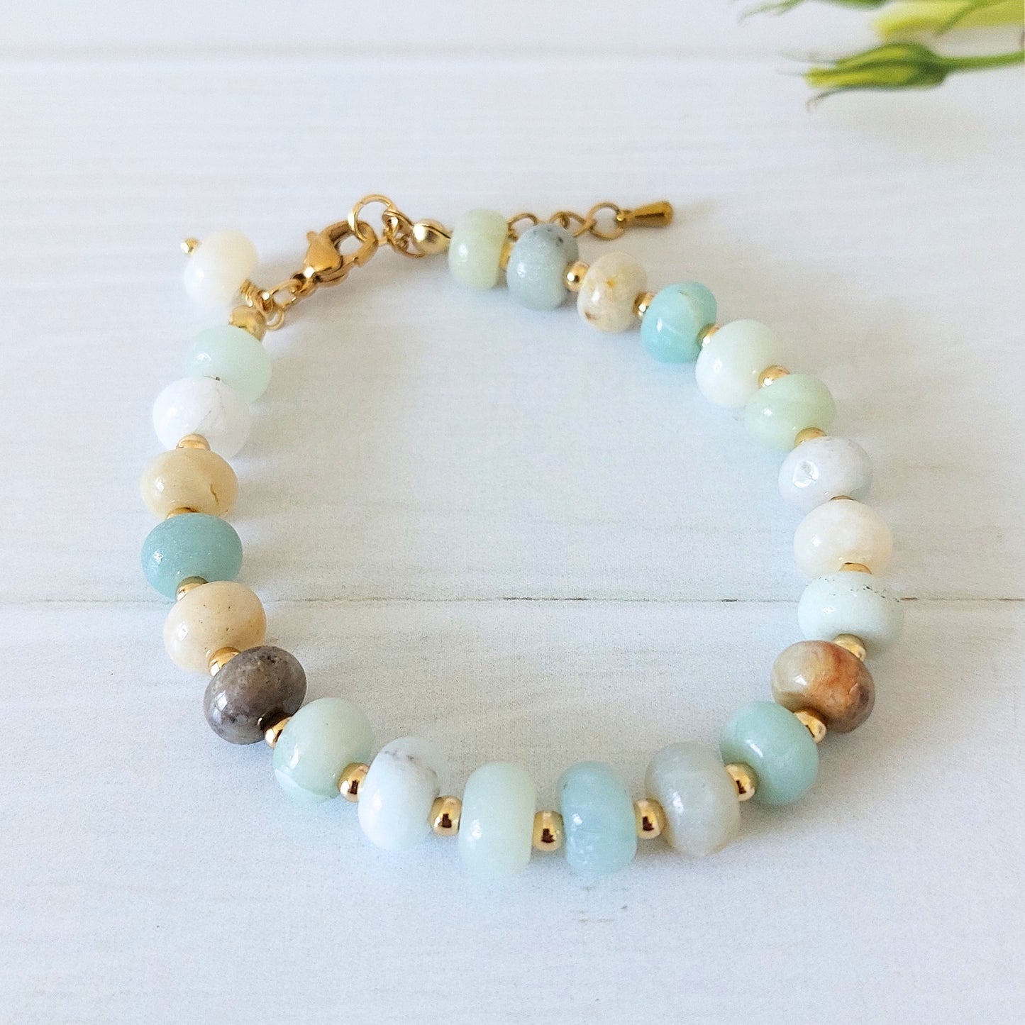 A beaded bracelet made of rondelle  gemstone beads in pale aqua, white, cream, and brown, separated by small gold-plated stainless steel spacer beads, with a gold clasp, photographed on a light wood surface.