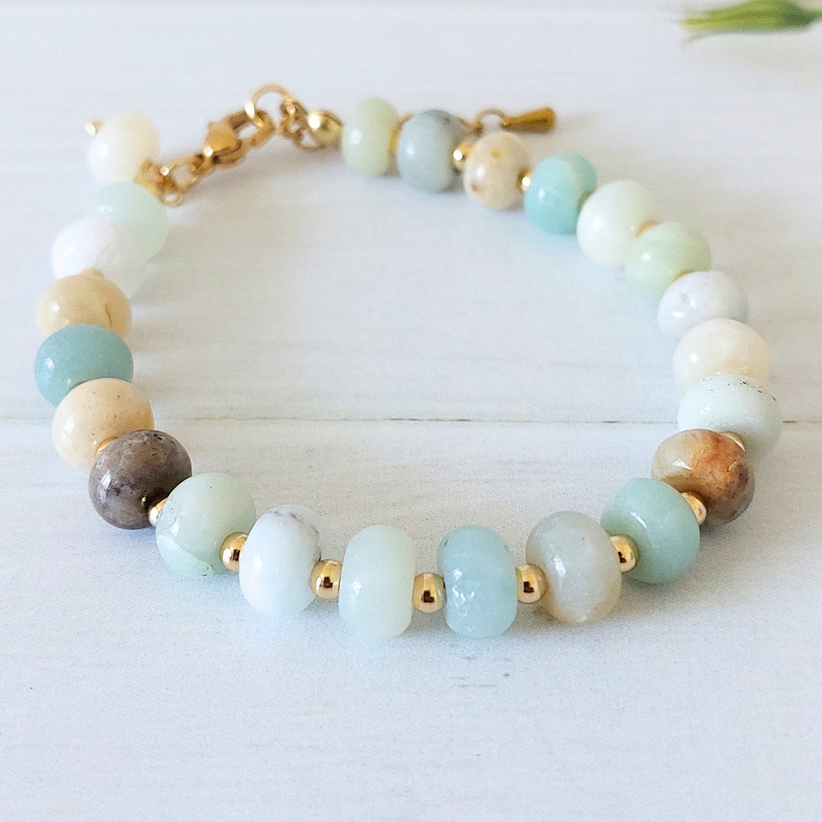 A beaded bracelet made of polished flower amazonite stones in pale blue, white, cream, and brown, separated by small gold-plated stainless steel spacer beads and joined with a gold clasp, photographed on a light wood surface.