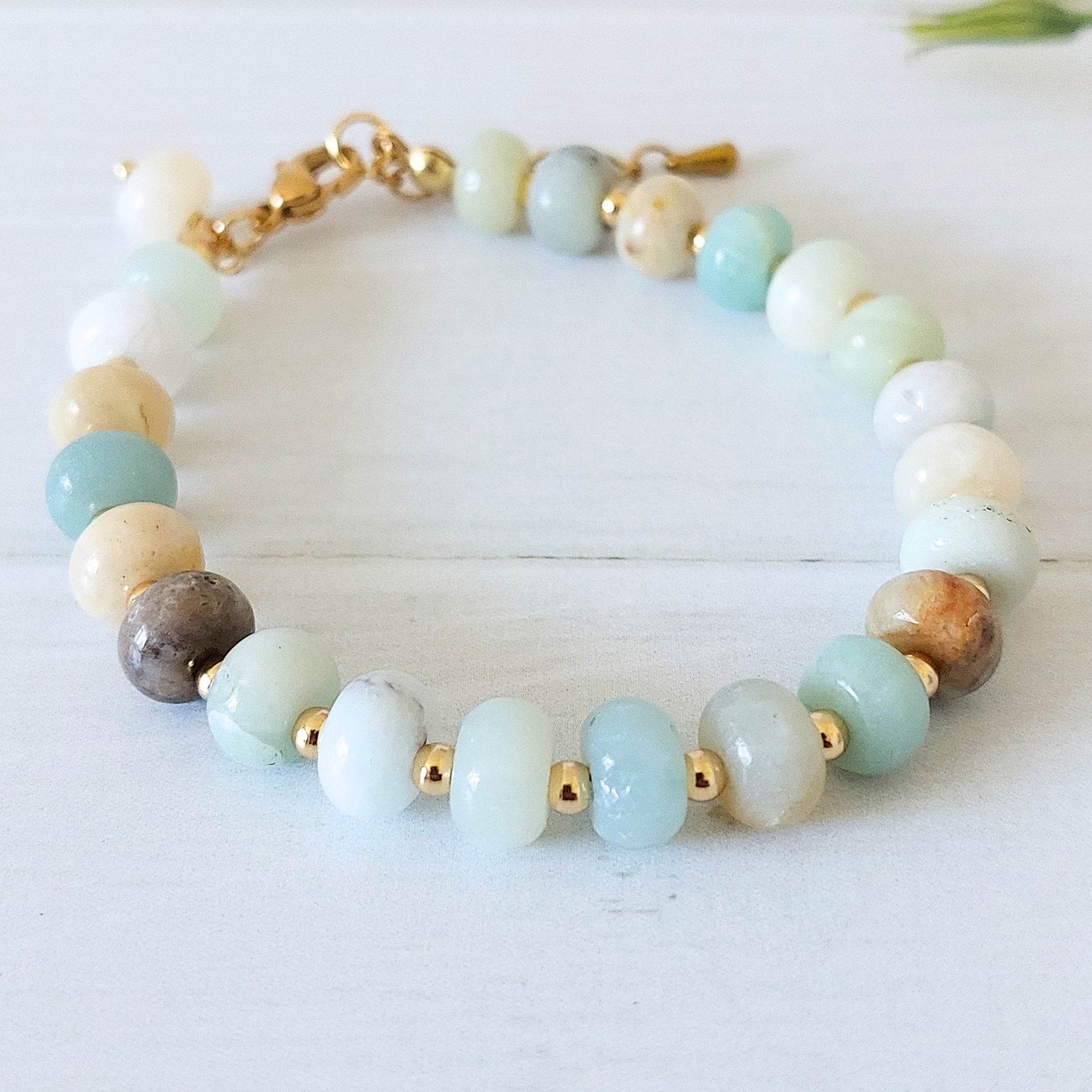 A beaded bracelet made of polished flower amazonite stones in pale blue, white, cream, and brown, separated by small gold-plated stainless steel spacer beads and joined with a gold clasp, photographed on a light wood surface.