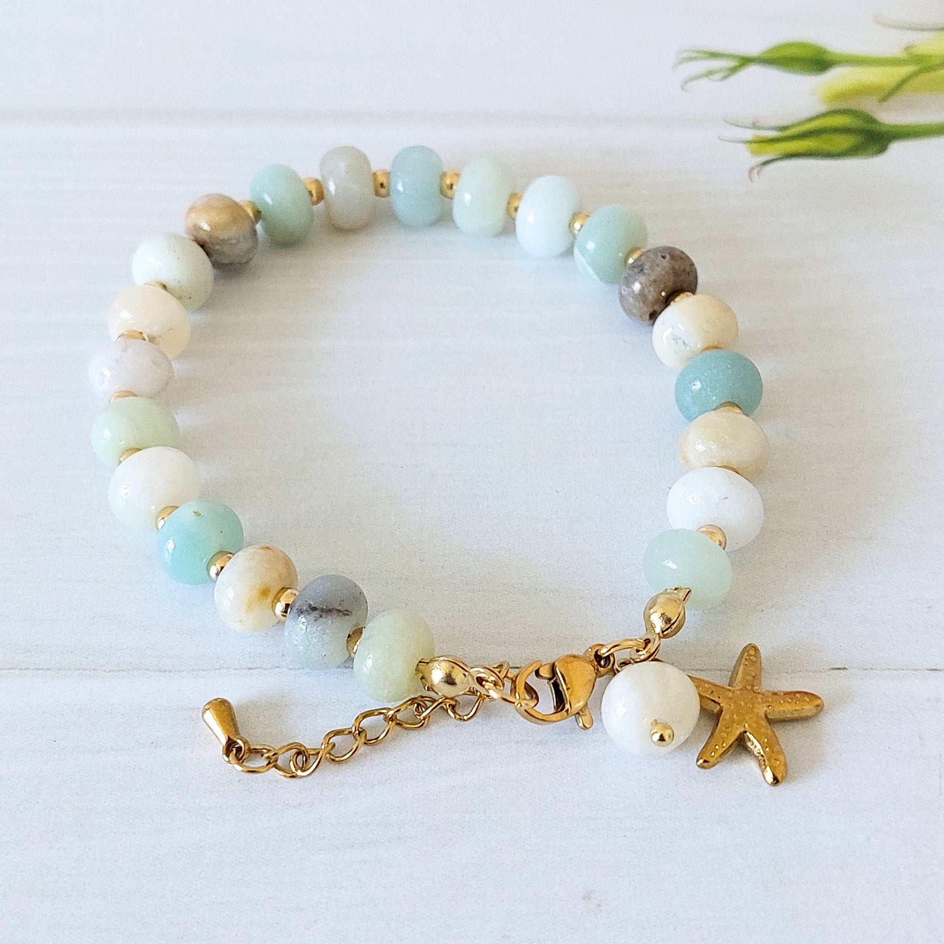 A beaded bracelet with light blue, mint green, white, and gray gemstone beads, accented by gold-plated stainless steel spacer beads, a gold clasp, an extender chain, a small white bead, and a gold starfish charm, displayed on a white wooden surface.