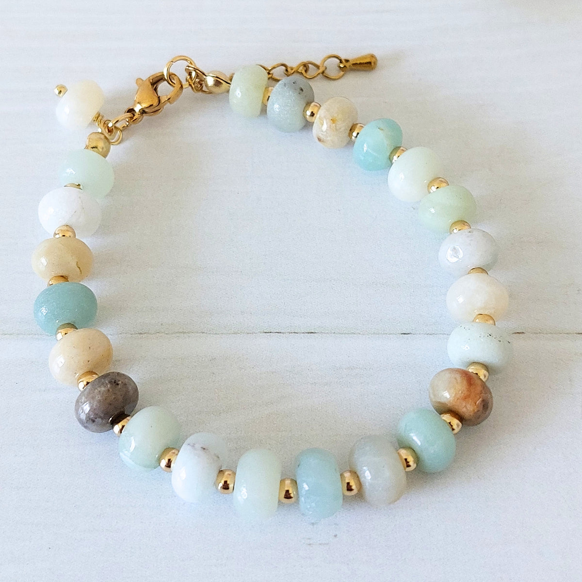 A beaded bracelet made of rondelle polished flower amazonite gemstone beads in light blue, white, cream, and brown, separated by small gold-plated stainless steel spacer beads and connected with a gold clasp and extender chain, displayed on a white wooden surface.