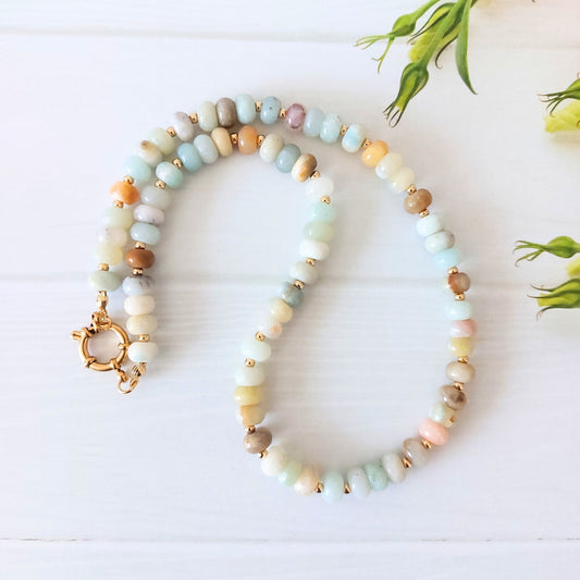 A beaded necklace made of pastel-colored flower amazonite rondelle beads in soft shades of blue, green, cream, pink, and brown, separated by small gold spacer beads and finished with a gold-plated stainless steel clasp, displayed on a white wooden background with green leaves in the corner.