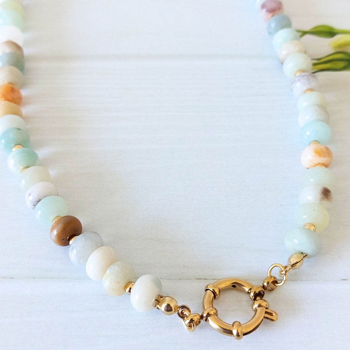 A beaded necklace made of  polished rondelle gemstone beads in soft pastel shades of mint, cream, white, and light brown, accented by a gold-plated stainless steel statement spring clasp, displayed on a white wooden surface.
