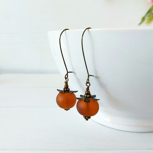 A pair of dangling earrings with frosted amber-orange glass beads and antique brass hooks, displayed against a white ceramic bowl on a light surface.