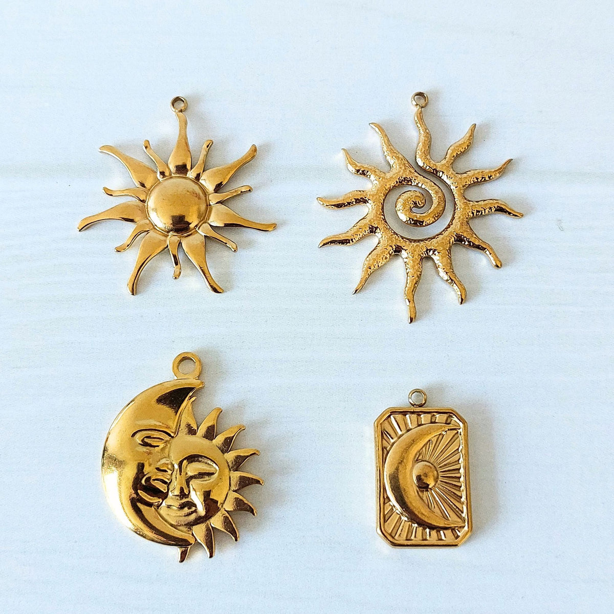 Four gold-toned celestial charms on a white background: two sun motifs, a crescent moon with a sun face, and a rectangular pendant with a crescent and sun design.