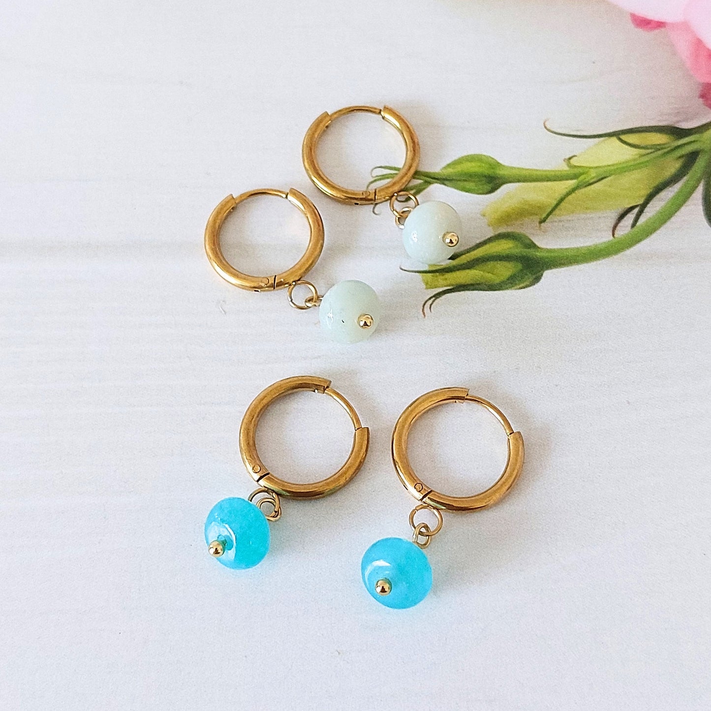 Four gold hoop earrings arranged on a white surface, with two featuring small pale blue flower amazonite bead drops and two featuring translucent turquoise-blue jade bead drops. A pink flower stem and green leaves are placed in the upper right corner as styling props.