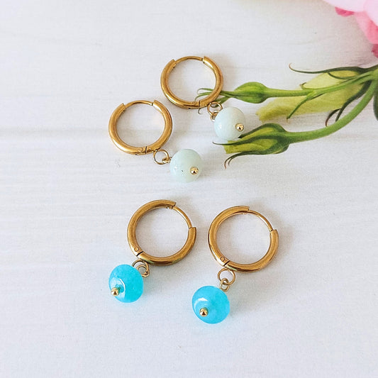 Four gold hoop earrings arranged on a white surface, with two featuring small pale blue flower amazonite bead drops and two featuring translucent turquoise-blue jade bead drops. A pink flower stem and green leaves are placed in the upper right corner as styling props.
