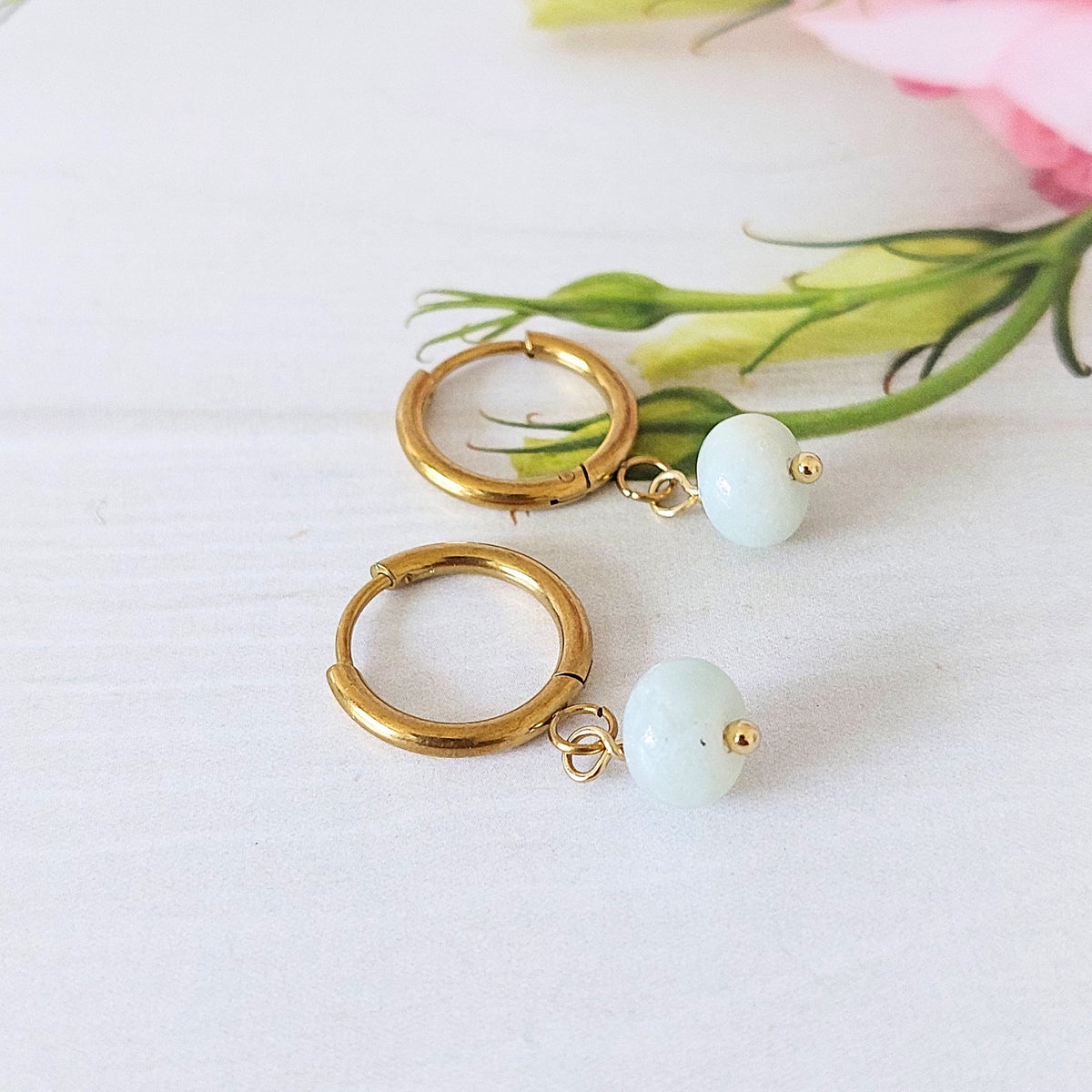 A pair of gold-plated stainless steel hoop earrings with a small light blue flower amazonite gemstone bead dangling from the end, displayed on a white surface with soft pink and green floral accents in the background.